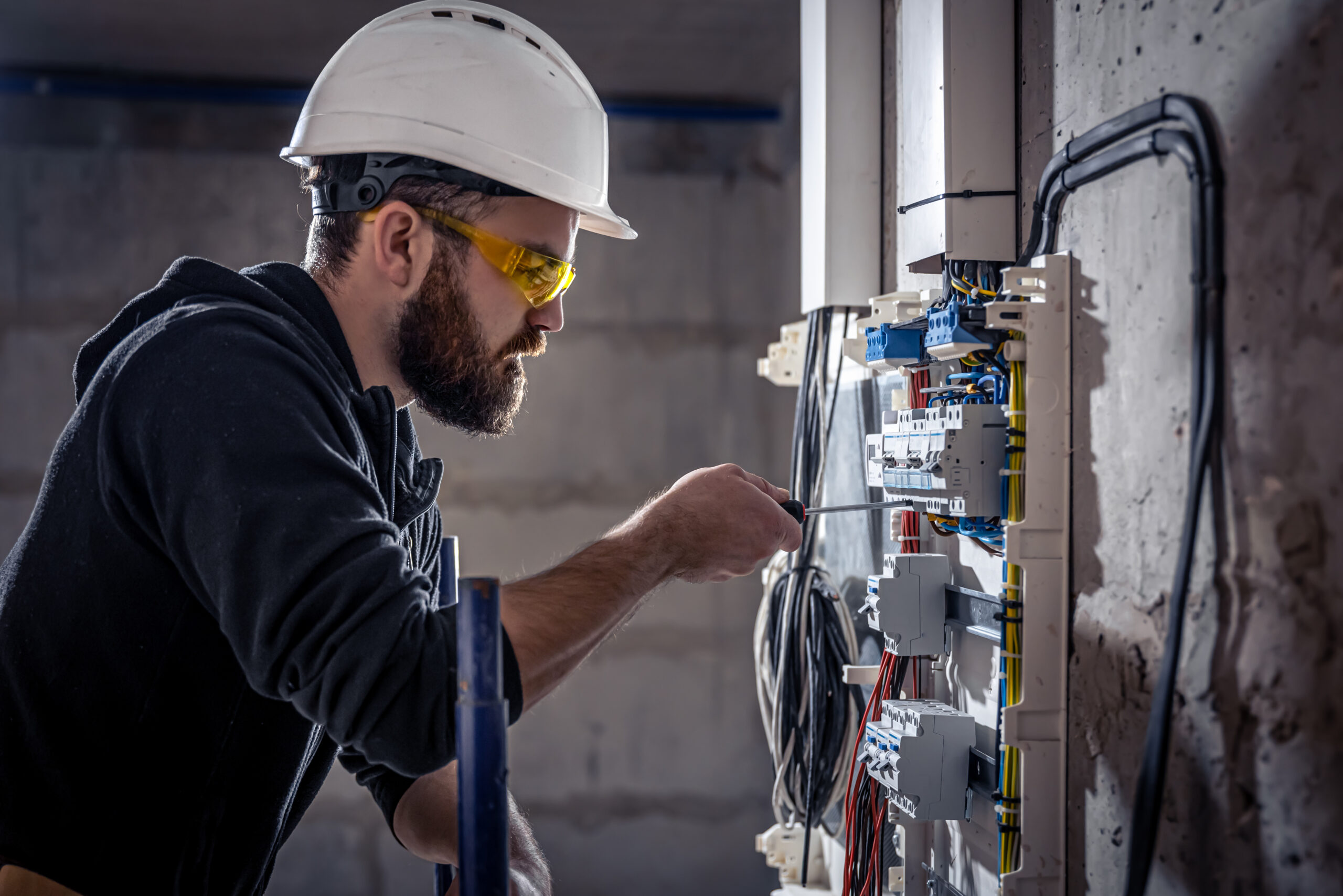 A male electrician works in a switchboard with an electrical connecting cable, connects the equipment with tools.